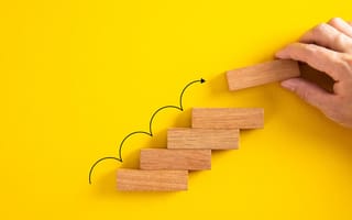 A hand stacks a rectangular wooden block on top of four other blocks to build a stairway, illustrating the cumulative nature of continuous learning.