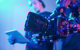 The foreground focuses on a video camera and an individual holding a notepad stands in the background.