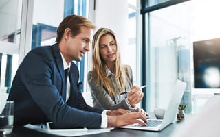 A pair of coworkers sit next each other behind a desk gesturing to a laptop screen.