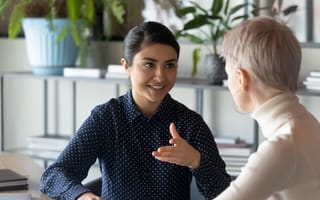 Friendly millennial businesswoman talking to blonde female colleague, sitting at table in office. Two young multiracial employees discussing working issues or enjoying informal conversation.