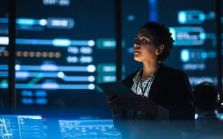 Young Female Government Employee Uses Tablet Computer in System Control Monitoring Center. In the Background Her Coworkers at Their Workspaces with Many Displays Showing Technical Data.