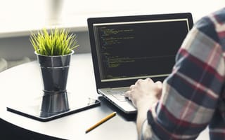 An over the shoulder view of a person typing on a laptop computer with lines of code on the screen.