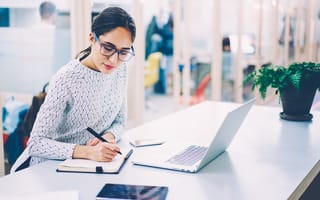 Dark-haired woman wearing glasses and red lipstick sits at a desk in front of a laptop. She takes written notes.