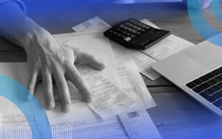 The hand of a person on a desk covered with papers, with a calculator in the background.
