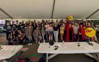 3Play team members are gathered around picnic tables under a big outdoor tent at a company event. They are wearing bright colors.