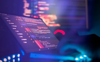 A close up of a pair of hands typing on a laptop keyboard with several lines of code on the screen to represent app development.
