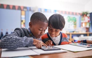 Two young students sit at a desk in a classroom looking closely at a tablet device.