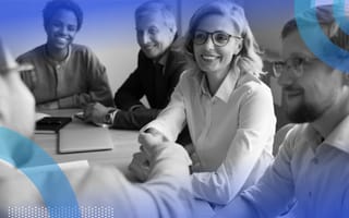 A group of colleagues seated around a table smiling while two of them shake hands.
