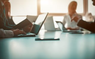 A board room table with laptops on it and people working on cpmputers