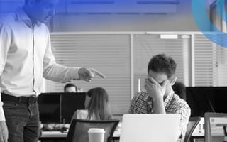 A person is sitting at a desk covering their eyes while a manager, who is standing, is pointing at them.