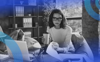 A young woman in glasses holding a coffee cup leafs through papers next to a laptop in an office full of books
