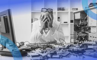 A frustrated man sitting at a desk littered with machine parts with his head in his hands and glasses on top of his head.