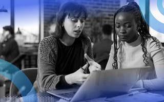Two women are sitting at a desk discussing something on a computer screen.
