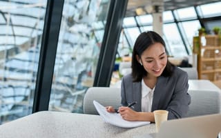 Woman sitting at table writing on paper while looking at her laptop.