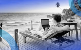 A person is sitting on a lounge chair with a laptop on a patio overlooking an ocean.