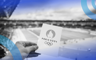 A hand is holding a card that says Paris 2024 in front of an athletic field.