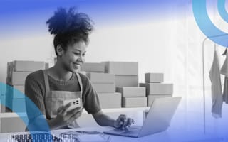A smiling small business owner sitting at a desk on her laptop with her smart phone in the other hand and stacks of boxes behind her.