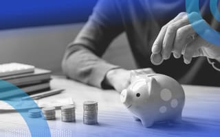 A person visible from the neck down and waist up sitting at a desk, adding a coin to a piggy bank next to inclining stacks of coins, dollar bills, a pen and a pile of notebooks.