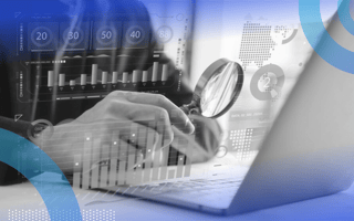 A businessman from the shoulders down and waist up sitting at a desk holding a magnifying glass to an open laptop with holographic symbols of data, numbers and charts overlayed on the image.