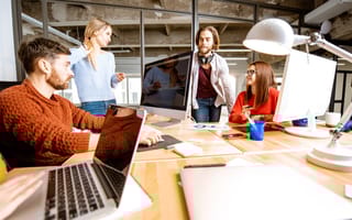 Team members chatting about project at table with desktop computers and laptops in office