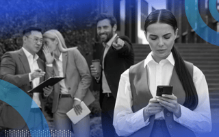 A woman standing and looking at her cell phone with a concerned expression while three colleagues stand behind her whispering and pointing at her.