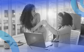 Two women colleagues, one sitting and one standing at a desk with an open laptop on it, fist bumping.