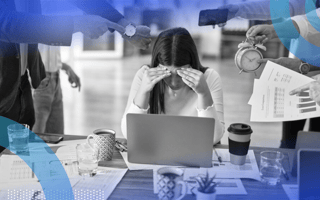 A woman hunched over her laptop shielding her face from several colleagues shoving clocks and papers at her.