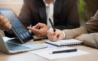 A close up of someone holding a tablet device with various data charts on the screen while another person writes in a notebook.
