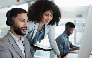 Colleagues looking at a monitor together, smiling. One is seated with a telephone headset, the other is standing next to him, leaning in.