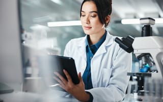 Woman scientist with dark hair in a lab coat, makes notes on a tablet. A microscope is visible in the foreground.