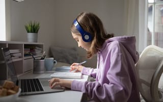 A young student sits at a desk wearing headphones and in front of a laptop while writing notes in a notebook.