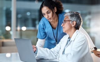 A medical professional in a white lab coat sits at a desk in front of a laptop computer while another professional wearing blue scrubs stands over their shoulder looking at the screen.