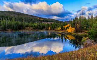 A blue sky with puffy white clouds reflected in a lake, surrounded by trees and a mountain peak.