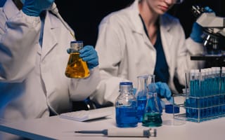 Two scientists wearing white lab coats and blue gloves sit at a desk handling test tubes and beakers filled with liquid.