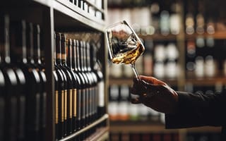 A close up of a hand holding a glass of white wine in front of a shelf filled with wine bottles.