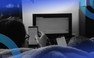 A view from behind of two people sitting on a couch, watching TV while also being on their smartphones.