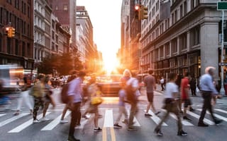 People are blurred across a city crosswalk, with the sun setting in the background.