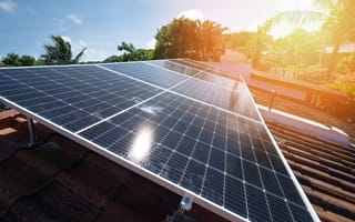 Solar panels sit on top of a roof with trees and sun shining in the background.
