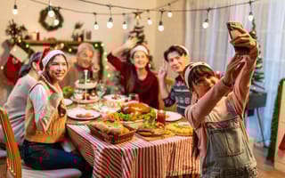 Family members gathered around a table filled with food and wearing festive attire pose for a group selfie while another family member stands at one end of the table to take the photo.