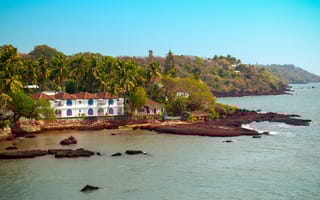 A large white resort-style building sits on Goa's coastline surrounded by trees next to the water.