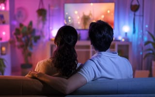 A couple is viewed from behind while cuddling on a sofa and watching television in a low-lit living room.