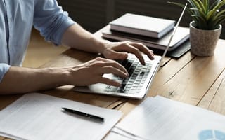 A close up of a pair of hands typing on a laptop keyboard sitting on a desk surrounded by books and papers.