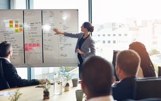 A woman leader points to an information on a whiteboard while giving a presentation in front of a group of colleagues in an office