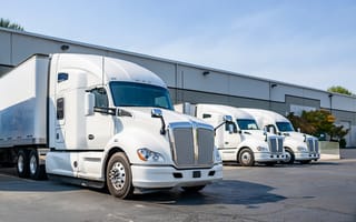 Three white cargo trucks are parked side-by-side in front of a warehouse.