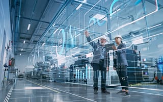 Two engineers in hard hats stand in a high-tech factory, engaging with virtual data displays projected in the air.