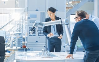 Three engineers in a modern robotics lab examine a robotic arm prototype. One woman holds a tablet and appears to be explaining a concept, while two men observe and discuss. The setting is high-tech, with lab equipment, CAD drawings, and advanced machiner