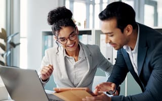 Two well-dressed professionals—a woman in a light gray blazer and glasses and a man in a dark suit—lean over a tablet together at a bright, modern office desk, with a laptop open nearby and shelves of binders in soft focus behind them.