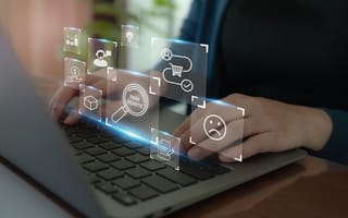 A close up of two hands typing on a laptop keyboard with icons hovering over it to represent customer research.