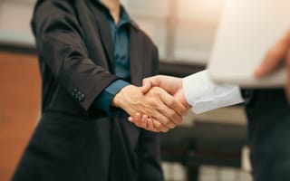 Two business professionals in formal attire shaking hands, symbolizing a business agreement or partnership. The people appear to be a man and a woman.