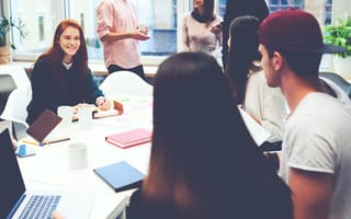 A group of young professionals collaborating in a brightly lit office space. One woman smiles while writing in a notebook at a table surrounded by laptops, notebooks, coffee mugs and sticky notes. Others are standing and chatting in the background near la